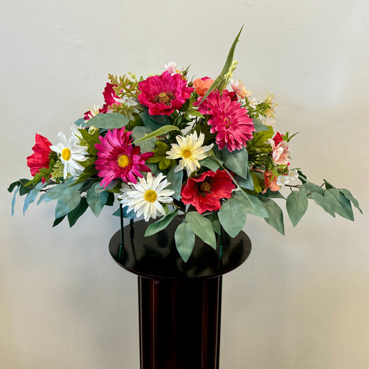 Introducing the Tombstone Saddle 60 cm Length by Posies Flowers & Fashion Inc., a vibrant floral arrangement featuring pink, red, and white flowers, including daisies and dahlias, complemented by lush green foliage. This versatile bouquet is elegantly displayed on a dark, cylindrical pedestal against a plain light gray background.