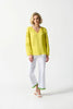 A person with short blonde hair is standing against a plain white background. They are wearing a bright yellow Georgette Puff Sleeve Top by Joseph Ribkoff, featuring long puff sleeves and a slit neckline, along with white pants adorned with green trim at the cuffs, and white open-toe shoes.