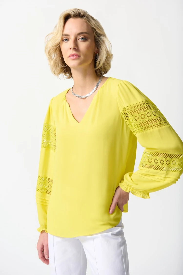 A person with short blonde hair is standing against a plain white background. They are wearing a bright yellow Georgette Puff Sleeve Top by Joseph Ribkoff, featuring long puff sleeves and a slit neckline, along with white pants adorned with green trim at the cuffs, and white open-toe shoes.