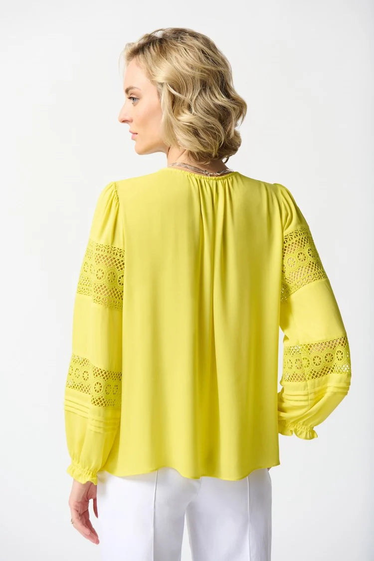 A person with short blonde hair is standing against a plain white background. They are wearing a bright yellow Georgette Puff Sleeve Top by Joseph Ribkoff, featuring long puff sleeves and a slit neckline, along with white pants adorned with green trim at the cuffs, and white open-toe shoes.