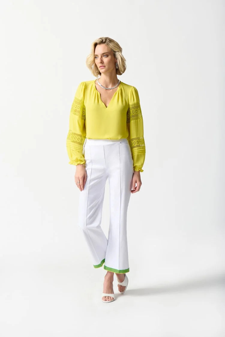 A person with short blonde hair is standing against a plain white background. They are wearing a bright yellow Georgette Puff Sleeve Top by Joseph Ribkoff, featuring long puff sleeves and a slit neckline, along with white pants adorned with green trim at the cuffs, and white open-toe shoes.