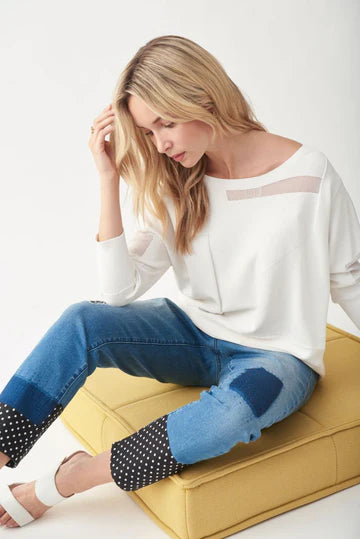 A woman with long blonde hair is sitting on a yellow ottoman, wearing the JOSEPH RIBKOFF TOP 221909 by Joseph Ribkoff, featuring a sheer panel at the neckline. She pairs it with denim jeans that have polka-dotted cuffs and patchwork details. Resting one hand on her chin, she looks directly at the camera.