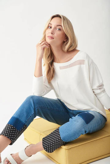 A woman with long blonde hair is sitting on a yellow ottoman, wearing the JOSEPH RIBKOFF TOP 221909 by Joseph Ribkoff, featuring a sheer panel at the neckline. She pairs it with denim jeans that have polka-dotted cuffs and patchwork details. Resting one hand on her chin, she looks directly at the camera.