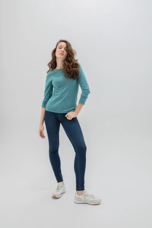 An individual with long, curly hair poses against a plain background. They are dressed in the high-quality Sweater 6377 MARBLE, featuring a long-sleeve design by MARBLE in an elegant teal shade. Paired with dark blue skinny jeans and white sneakers, the person strikes a confident pose with their left leg slightly bent, their left hand resting on their hip while their right arm hangs loosely by their side.