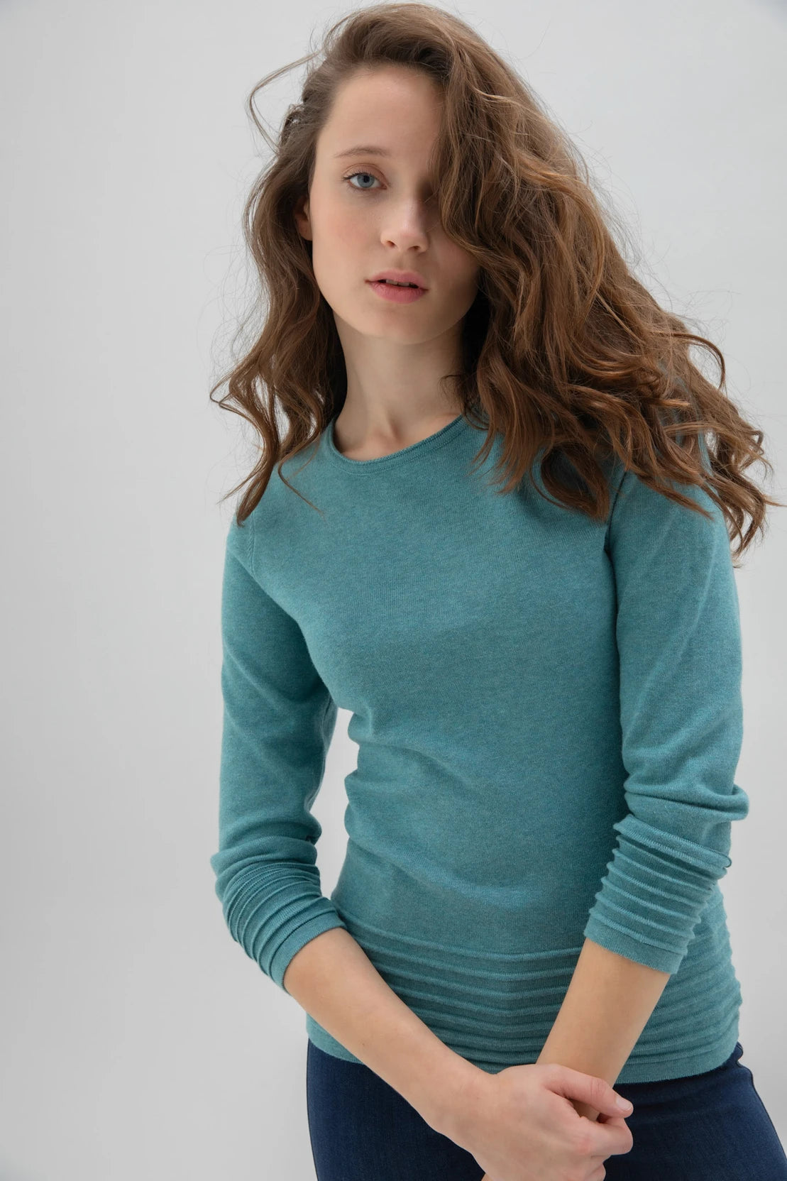 An individual with long, curly hair poses against a plain background. They are dressed in the high-quality Sweater 6377 MARBLE, featuring a long-sleeve design by MARBLE in an elegant teal shade. Paired with dark blue skinny jeans and white sneakers, the person strikes a confident pose with their left leg slightly bent, their left hand resting on their hip while their right arm hangs loosely by their side.