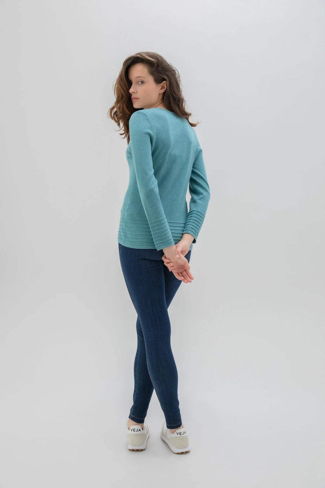 An individual with long, curly hair poses against a plain background. They are dressed in the high-quality Sweater 6377 MARBLE, featuring a long-sleeve design by MARBLE in an elegant teal shade. Paired with dark blue skinny jeans and white sneakers, the person strikes a confident pose with their left leg slightly bent, their left hand resting on their hip while their right arm hangs loosely by their side.