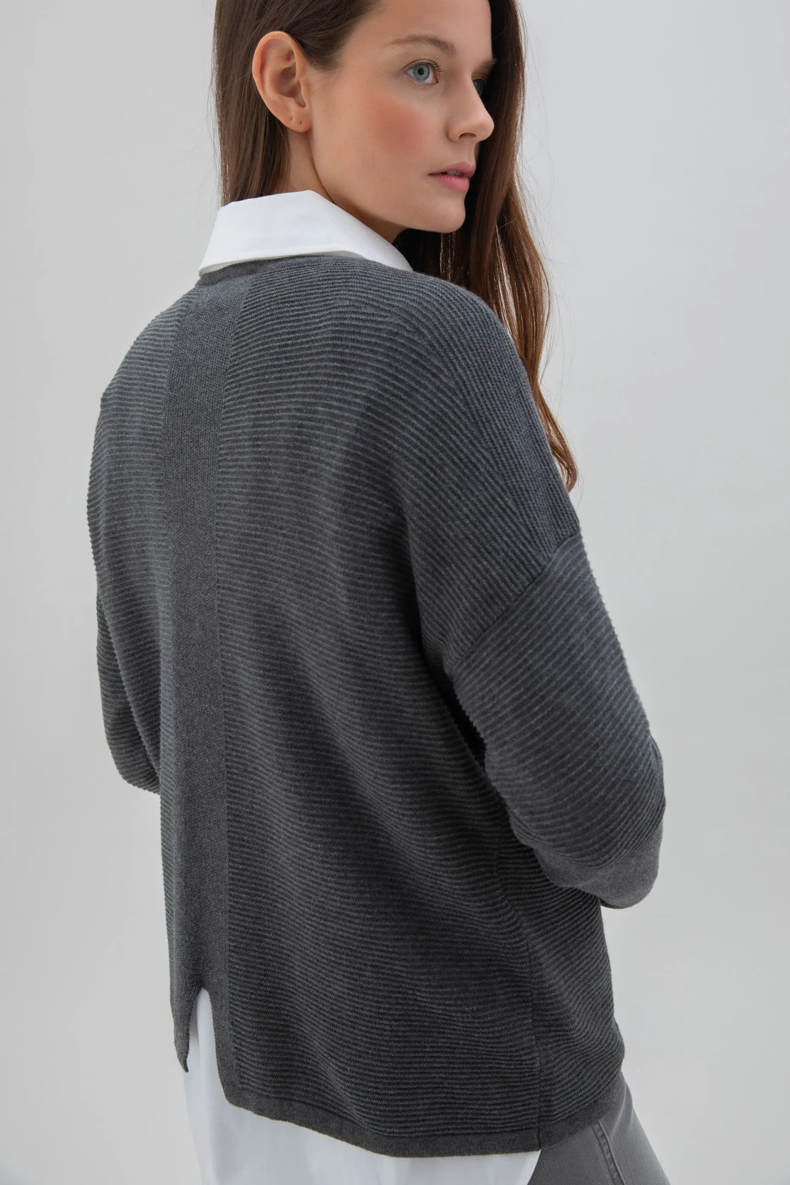 A woman with long brown hair is posing against a plain background. She is dressed in the Sweater 7553-105 MARBLE, a high-quality dark gray sweater from the Marble brand, along with a white shirt, light gray leggings, and white sneakers. Her expression is neutral as she stands with one hand by her side and the other slightly bent.