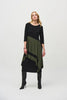 A woman stands against a plain background, wearing the Joseph Ribkoff Silky Knit Colour Block Handkerchief Dress 244186 in black and olive green. The asymmetrical dress, made from a silky knit fabric, features three-quarter sleeves and a handkerchief hemline that ends at mid-calf length. She completes her outfit with black knee-high boots. Her expression is neutral, and her hands rest at her sides.