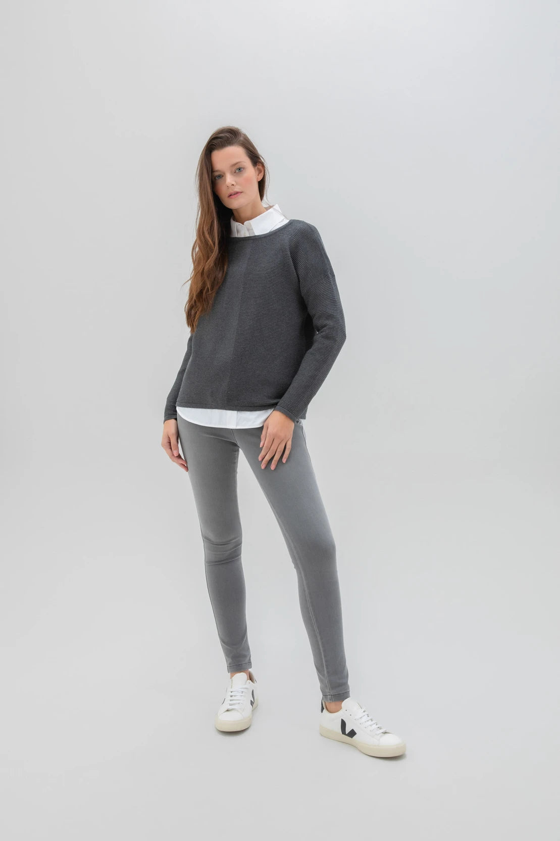 A woman with long brown hair is posing against a plain background. She is dressed in the Sweater 7553-105 MARBLE, a high-quality dark gray sweater from the Marble brand, along with a white shirt, light gray leggings, and white sneakers. Her expression is neutral as she stands with one hand by her side and the other slightly bent.