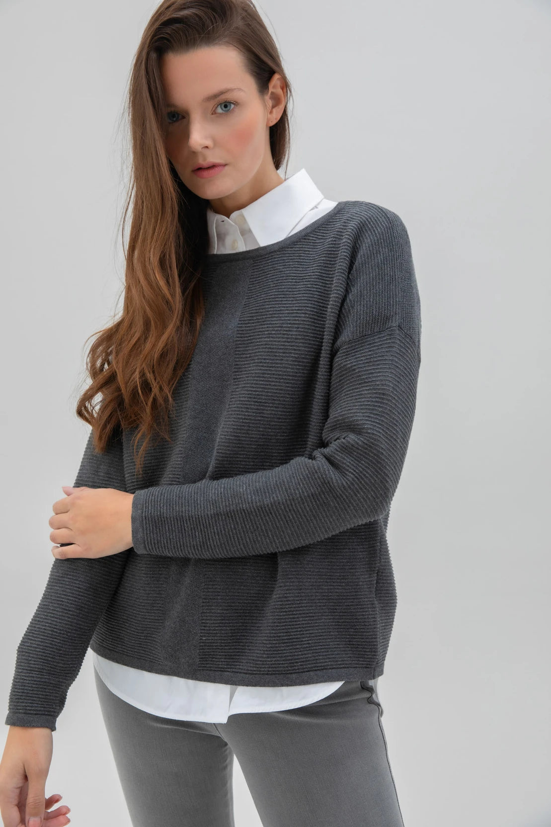 A woman with long brown hair is posing against a plain background. She is dressed in the Sweater 7553-105 MARBLE, a high-quality dark gray sweater from the Marble brand, along with a white shirt, light gray leggings, and white sneakers. Her expression is neutral as she stands with one hand by her side and the other slightly bent.