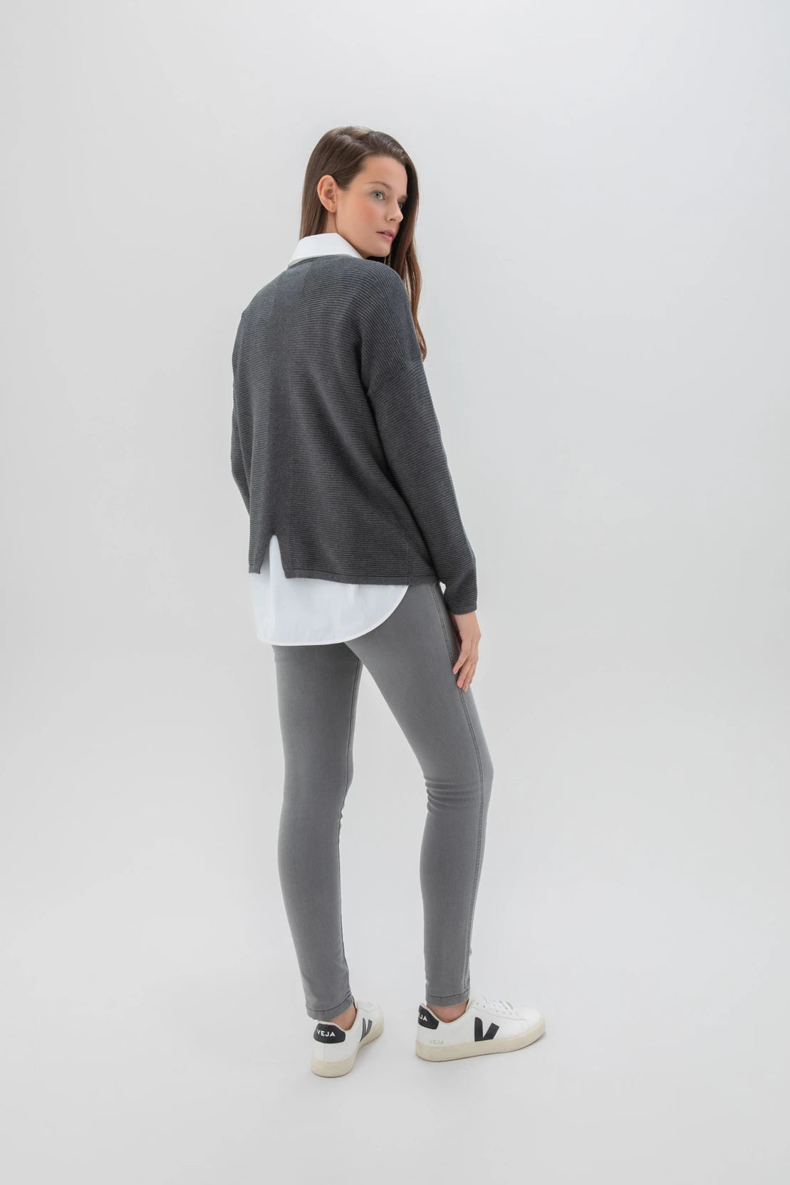 A woman with long brown hair is posing against a plain background. She is dressed in the Sweater 7553-105 MARBLE, a high-quality dark gray sweater from the Marble brand, along with a white shirt, light gray leggings, and white sneakers. Her expression is neutral as she stands with one hand by her side and the other slightly bent.