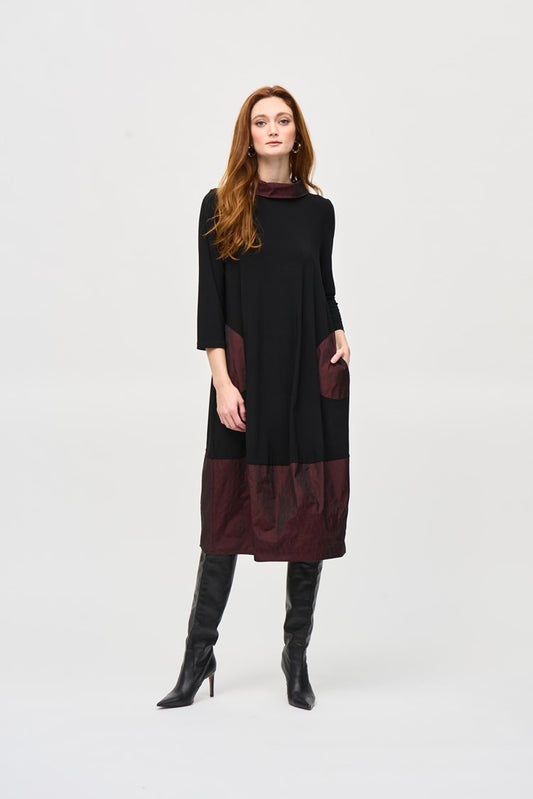 A woman with long red hair stands against a plain background, wearing the Silky Knit And Memory Cocoon Dress 243080 by Joseph Ribkoff. The dress features dark red accents on the pockets and hem, which she pairs with black knee-high boots. She has a confident expression and her hands in her dress pockets.