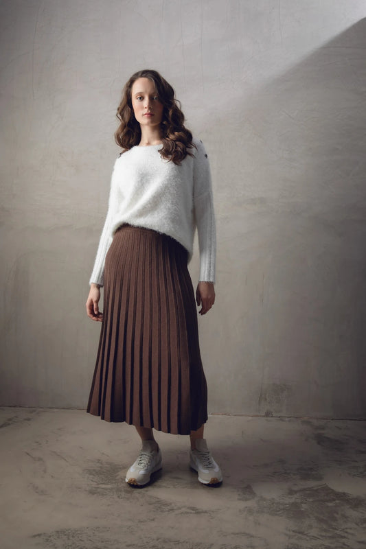 A woman stands against a textured wall, wearing the Sweater 7545-104 MARBLE from the brand MARBLE and a brown pleated midi skirt. She also has on white sneakers and appears relaxed, with loose, wavy hair cascading over her shoulders. The natural light illuminating her is reminiscent of the soft glow over marble in Scotland.