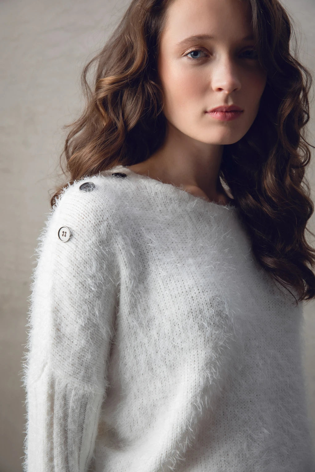 A woman stands against a textured wall, wearing the Sweater 7545-104 MARBLE from the brand MARBLE and a brown pleated midi skirt. She also has on white sneakers and appears relaxed, with loose, wavy hair cascading over her shoulders. The natural light illuminating her is reminiscent of the soft glow over marble in Scotland.