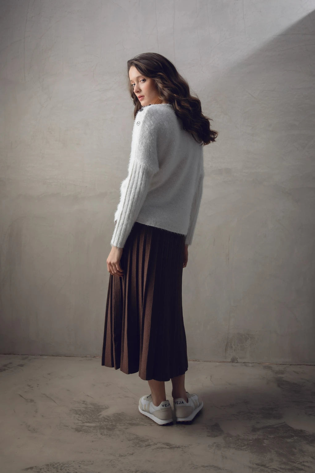 A woman stands against a textured wall, wearing the Sweater 7545-104 MARBLE from the brand MARBLE and a brown pleated midi skirt. She also has on white sneakers and appears relaxed, with loose, wavy hair cascading over her shoulders. The natural light illuminating her is reminiscent of the soft glow over marble in Scotland.