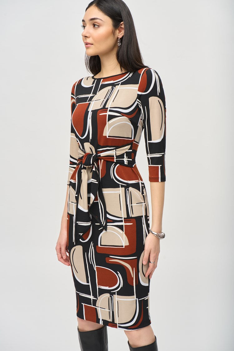 A woman is standing and wearing the Silky Knit Abstract Puff Print Sheath Dress 243325 by Joseph Ribkoff. The knee-length dress features an abstract puff print in shades of black, white, brown, and beige. The boat neckline adds elegance while the dress is cinched at the waist with a matching fabric belt. She has long, dark hair and accessorizes with earrings and a bracelet.