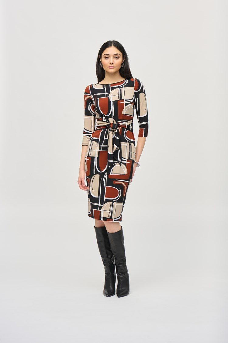 A woman is standing and wearing the Silky Knit Abstract Puff Print Sheath Dress 243325 by Joseph Ribkoff. The knee-length dress features an abstract puff print in shades of black, white, brown, and beige. The boat neckline adds elegance while the dress is cinched at the waist with a matching fabric belt. She has long, dark hair and accessorizes with earrings and a bracelet.