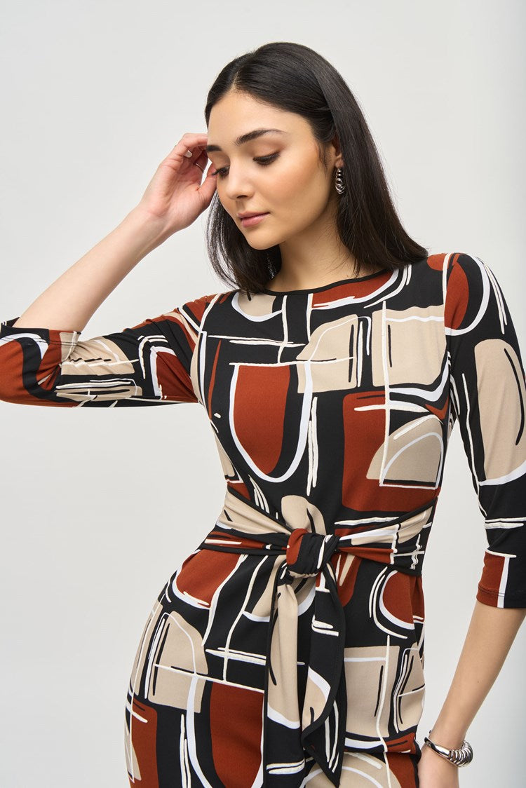 A woman is standing and wearing the Silky Knit Abstract Puff Print Sheath Dress 243325 by Joseph Ribkoff. The knee-length dress features an abstract puff print in shades of black, white, brown, and beige. The boat neckline adds elegance while the dress is cinched at the waist with a matching fabric belt. She has long, dark hair and accessorizes with earrings and a bracelet.