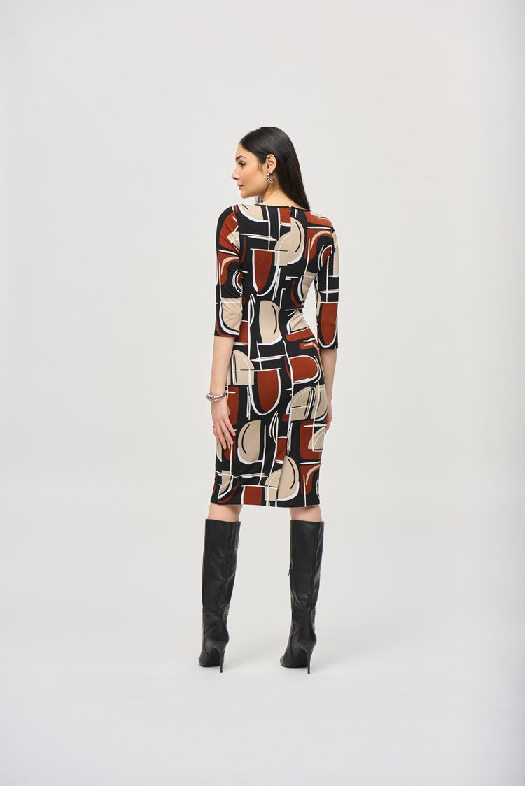 A woman is standing and wearing the Silky Knit Abstract Puff Print Sheath Dress 243325 by Joseph Ribkoff. The knee-length dress features an abstract puff print in shades of black, white, brown, and beige. The boat neckline adds elegance while the dress is cinched at the waist with a matching fabric belt. She has long, dark hair and accessorizes with earrings and a bracelet.