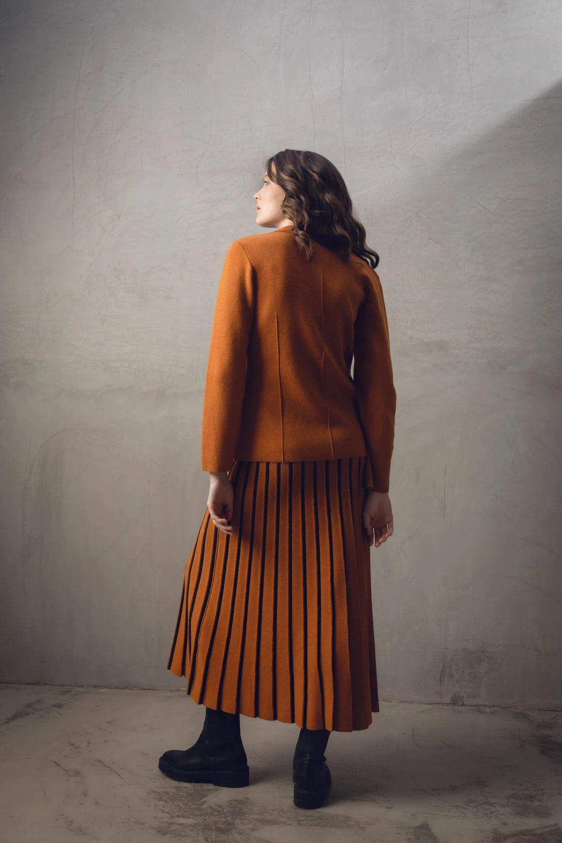 A woman with wavy brown hair stands with her back to the camera, showcasing a stylish burnt orange Sweater 7597-224 MARBLE and matching pleated skirt paired with black boots. The background is a neutral-toned textured wall.