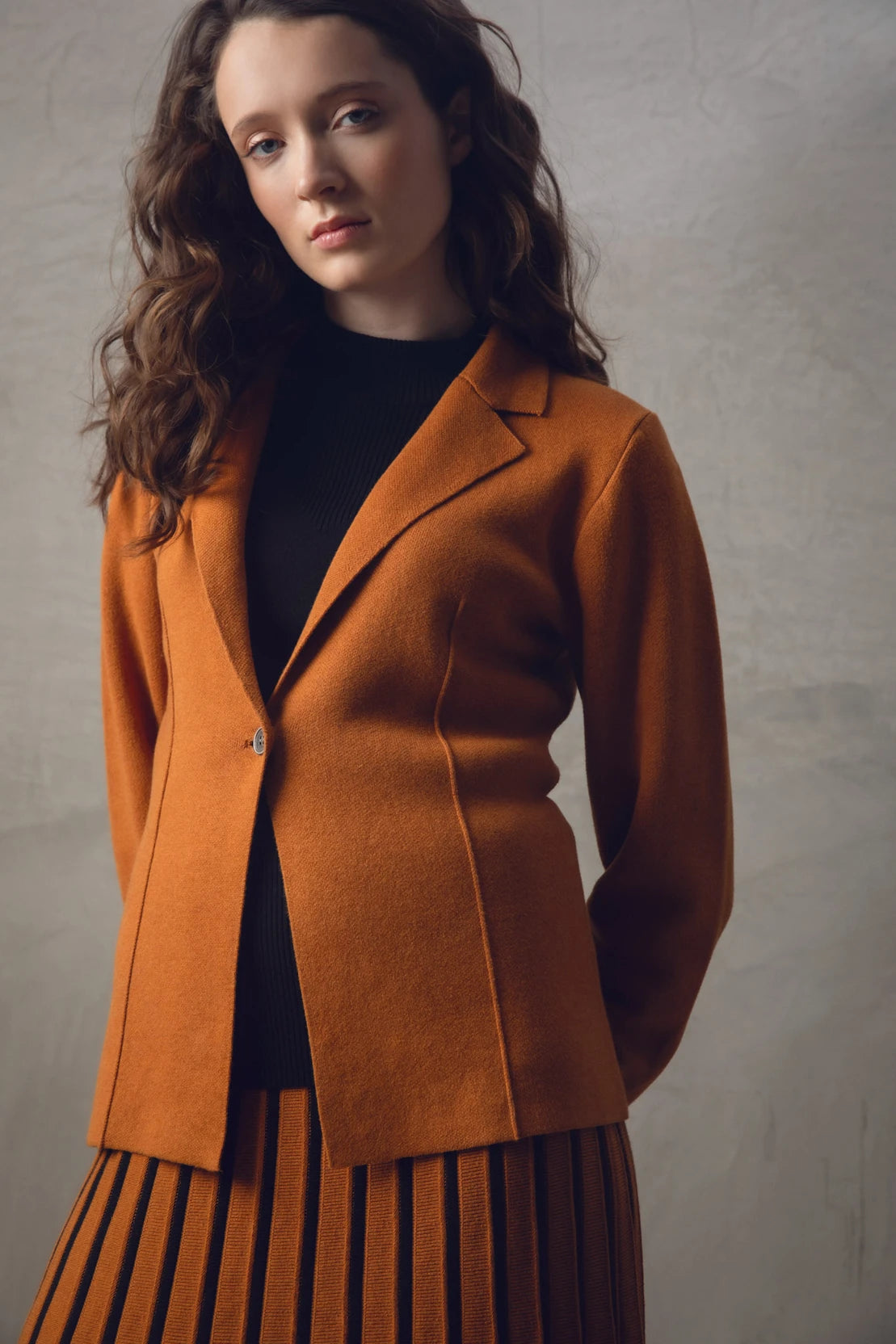 A woman with wavy brown hair stands with her back to the camera, showcasing a stylish burnt orange Sweater 7597-224 MARBLE and matching pleated skirt paired with black boots. The background is a neutral-toned textured wall.