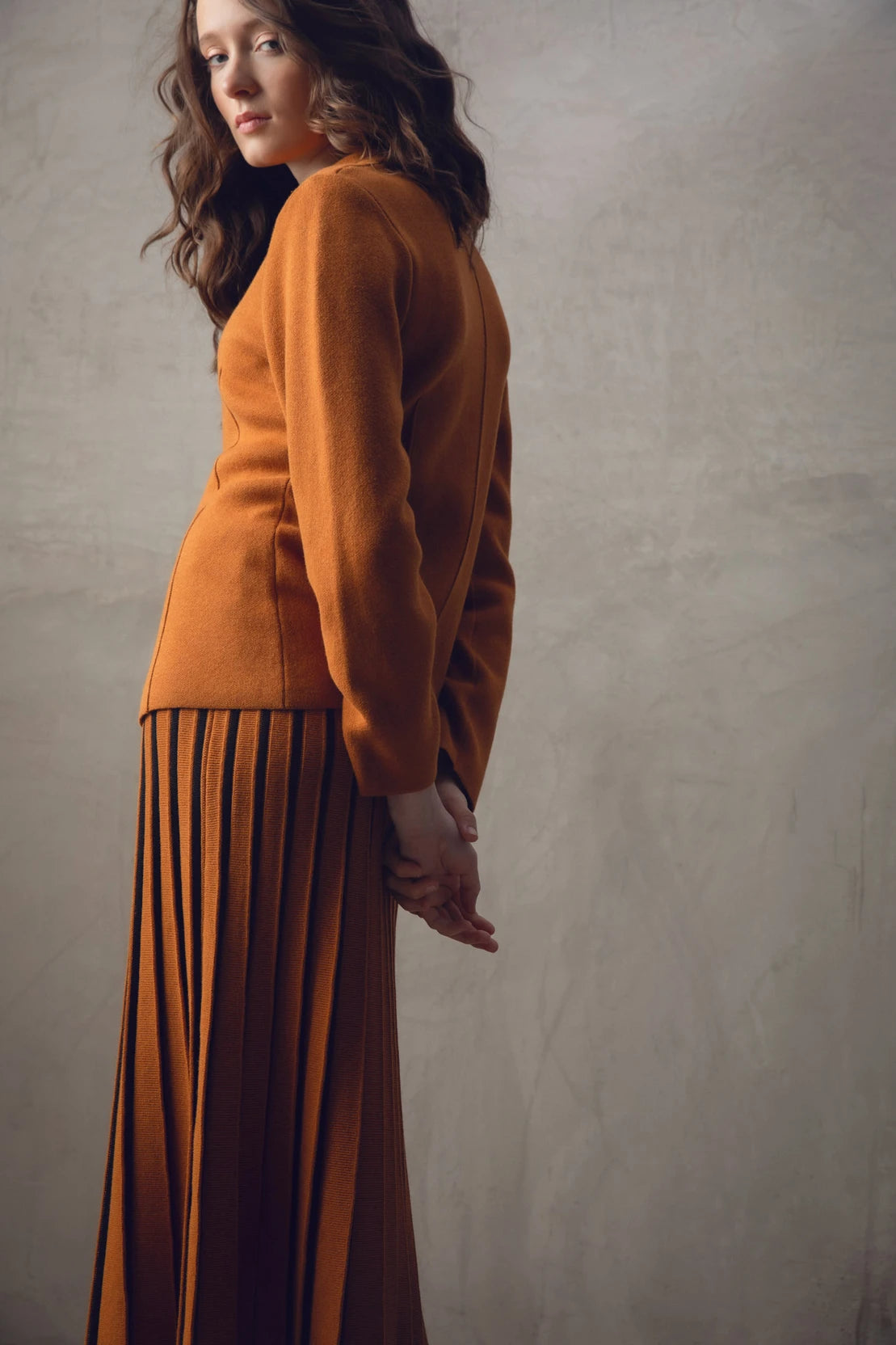 A woman with wavy brown hair stands with her back to the camera, showcasing a stylish burnt orange Sweater 7597-224 MARBLE and matching pleated skirt paired with black boots. The background is a neutral-toned textured wall.