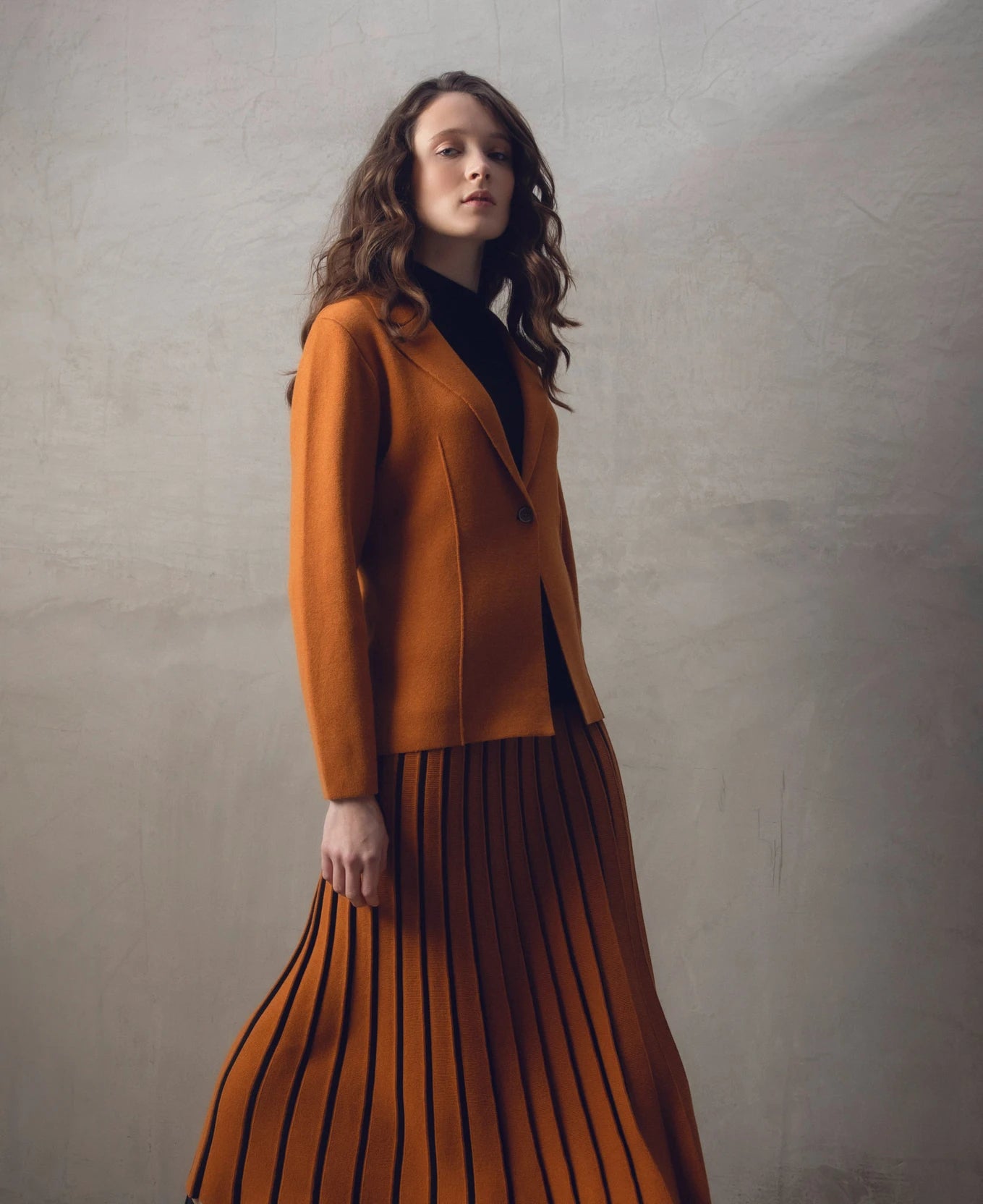 A woman with wavy brown hair stands with her back to the camera, showcasing a stylish burnt orange Sweater 7597-224 MARBLE and matching pleated skirt paired with black boots. The background is a neutral-toned textured wall.