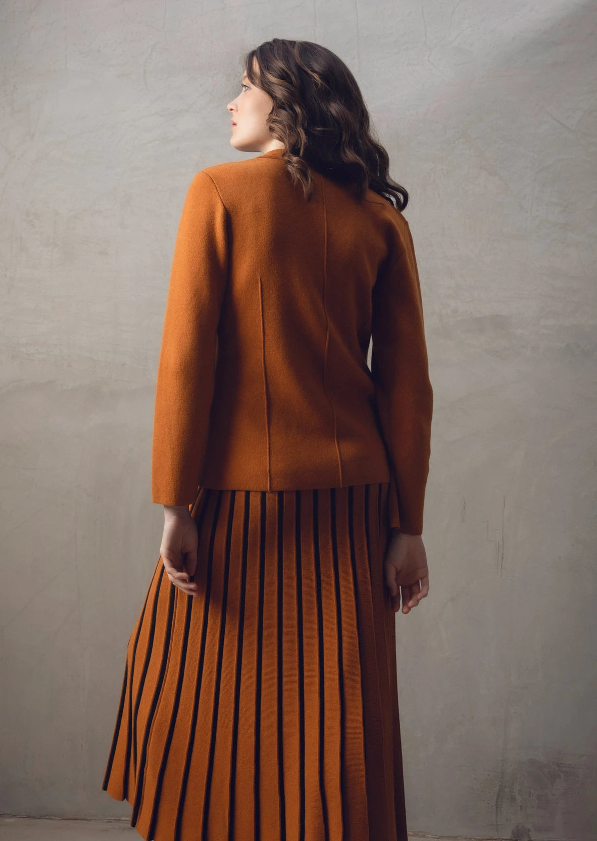 A woman with wavy brown hair stands with her back to the camera, showcasing a stylish burnt orange Sweater 7597-224 MARBLE and matching pleated skirt paired with black boots. The background is a neutral-toned textured wall.