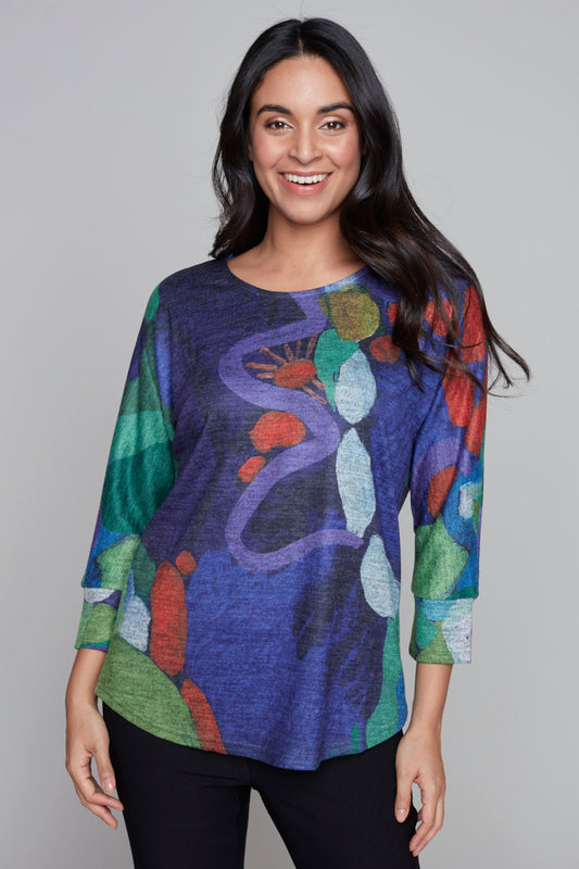 A person with long hair is smiling at the camera. They are wearing a vibrant Knit Top 91703 by Claire Desjardins, showcasing an abstract pattern in hues of blue, green, red, and white. The stylish top features long sleeves, and the individual is standing against a plain gray background.