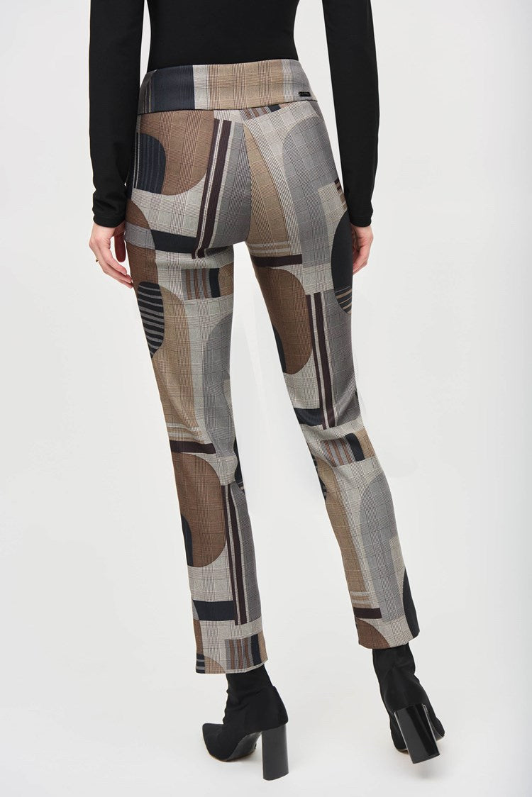 A woman stands against a plain white background, wearing a black wrap-style top with long sleeves and Jacquard Abstract Print Straight Pull-On Pants 243299 by Joseph Ribkoff, featuring abstract patterns in shades of brown, gray, and black. She completes the outfit with black ankle boots. She has long, reddish-brown hair and a neutral expression.