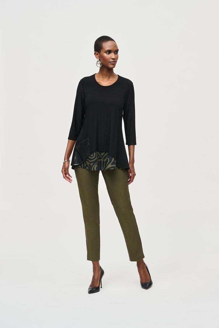 A woman stands confidently, wearing a black three-quarter sleeve blouse with patterned details at the hem and Classic Slim Pant - Seasonal Colors 201483F24 Joseph Ribkoff in olive green. She pairs the outfit with black high-heels and hoop earrings. The background is plain white.