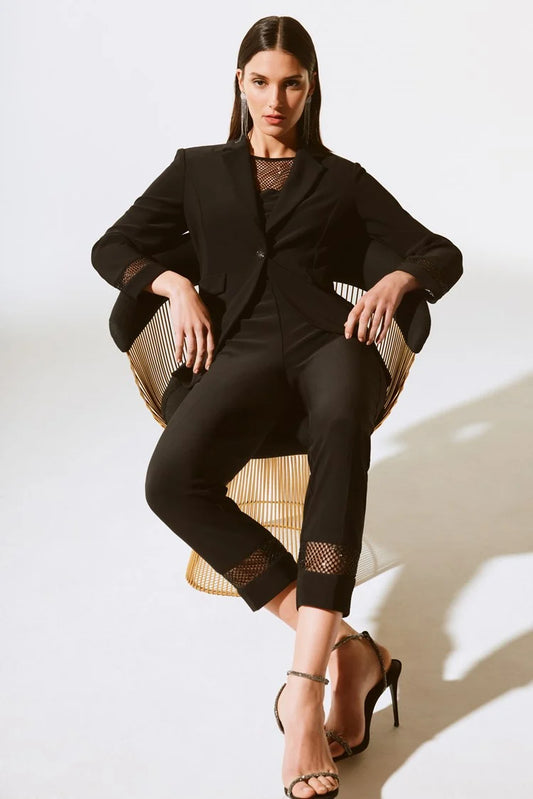 A woman with long dark hair is seated in a stylish wireframe chair. She is dressed in a black suit from the Joseph Ribkoff collection, specifically the Silky Knit And Rhinestone Mesh Pants 243753 Joseph Ribkoff, paired with black stiletto heels. The white background and her confident pose create a sophisticated and modern look.