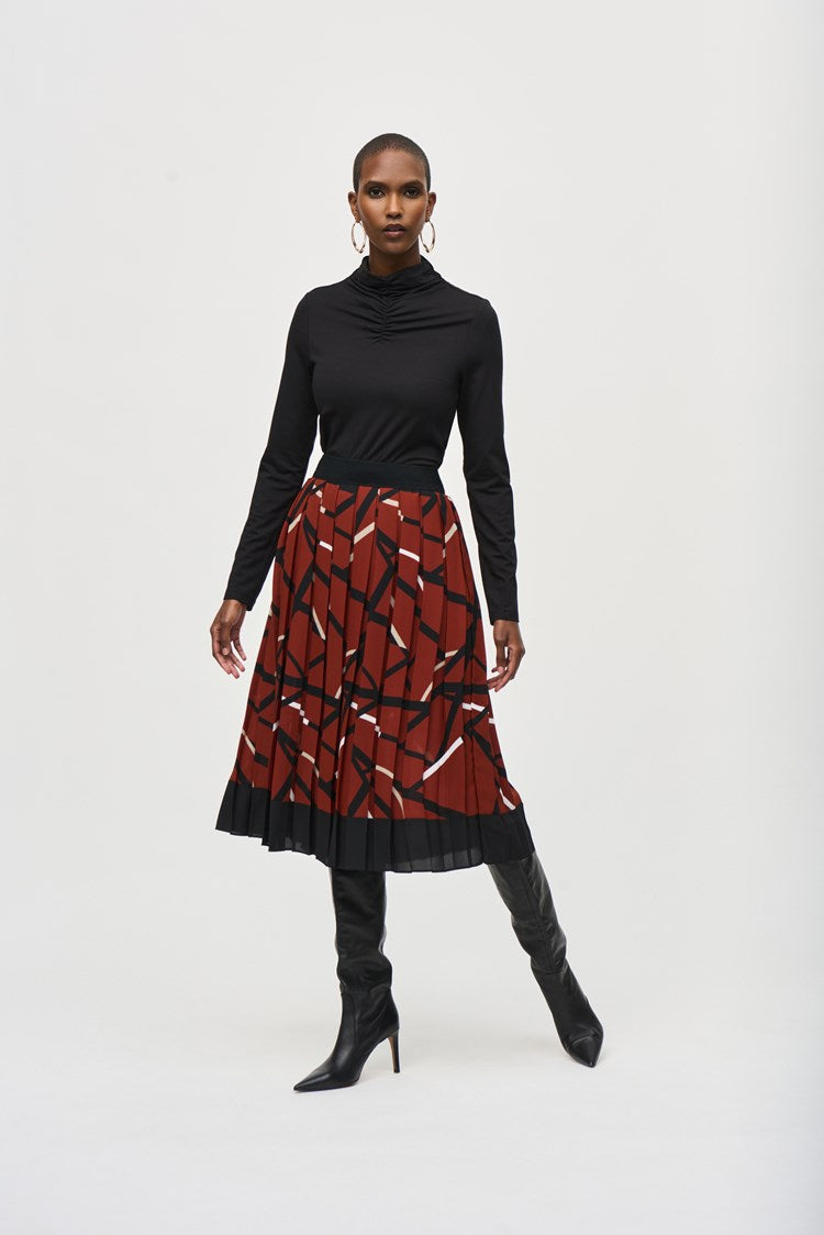 A woman stands confidently against a plain white background, wearing a black long-sleeved top and a Joseph Ribkoff Georgette Geometric Print Pleated Skirt 243201. She pairs the outfit with high-heeled black boots and large hoop earrings. Her hands are relaxed by her sides.