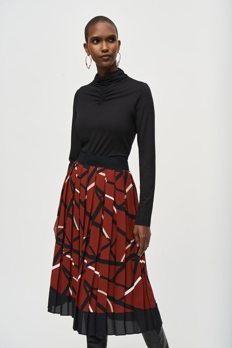 A woman stands confidently against a plain white background, wearing a black long-sleeved top and a Joseph Ribkoff Georgette Geometric Print Pleated Skirt 243201. She pairs the outfit with high-heeled black boots and large hoop earrings. Her hands are relaxed by her sides.