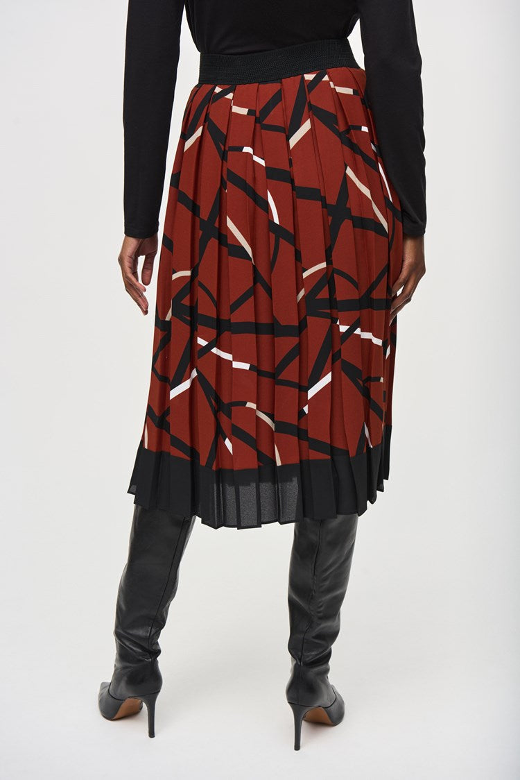 A woman stands confidently against a plain white background, wearing a black long-sleeved top and a Joseph Ribkoff Georgette Geometric Print Pleated Skirt 243201. She pairs the outfit with high-heeled black boots and large hoop earrings. Her hands are relaxed by her sides.