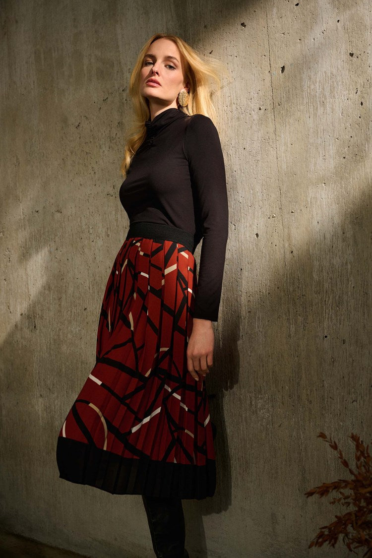 A woman stands confidently against a plain white background, wearing a black long-sleeved top and a Joseph Ribkoff Georgette Geometric Print Pleated Skirt 243201. She pairs the outfit with high-heeled black boots and large hoop earrings. Her hands are relaxed by her sides.