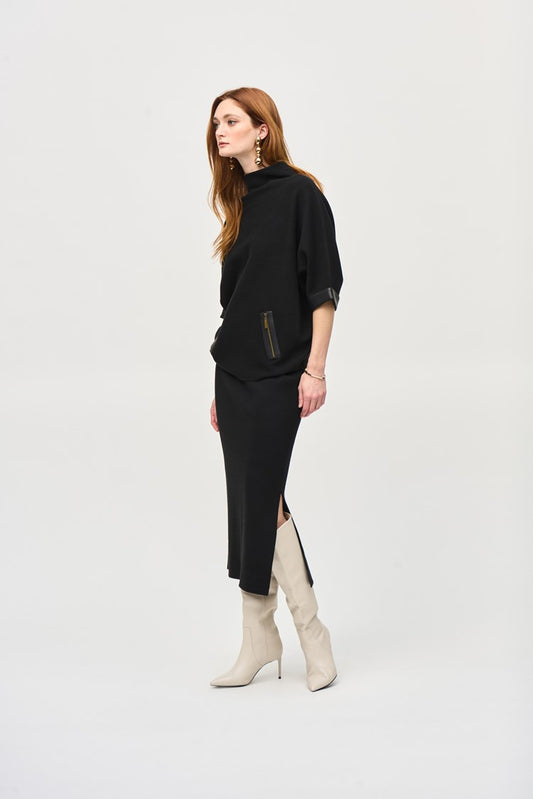 A person with long, red hair is standing against a plain white background. They are wearing a black, loose-fitting top with pocket details and the Joseph Ribkoff Sweater Knit Midi Skirt (style 243967) featuring a side slit and covered elastic waistband. They are also wearing white, heeled boots and dangling earrings.