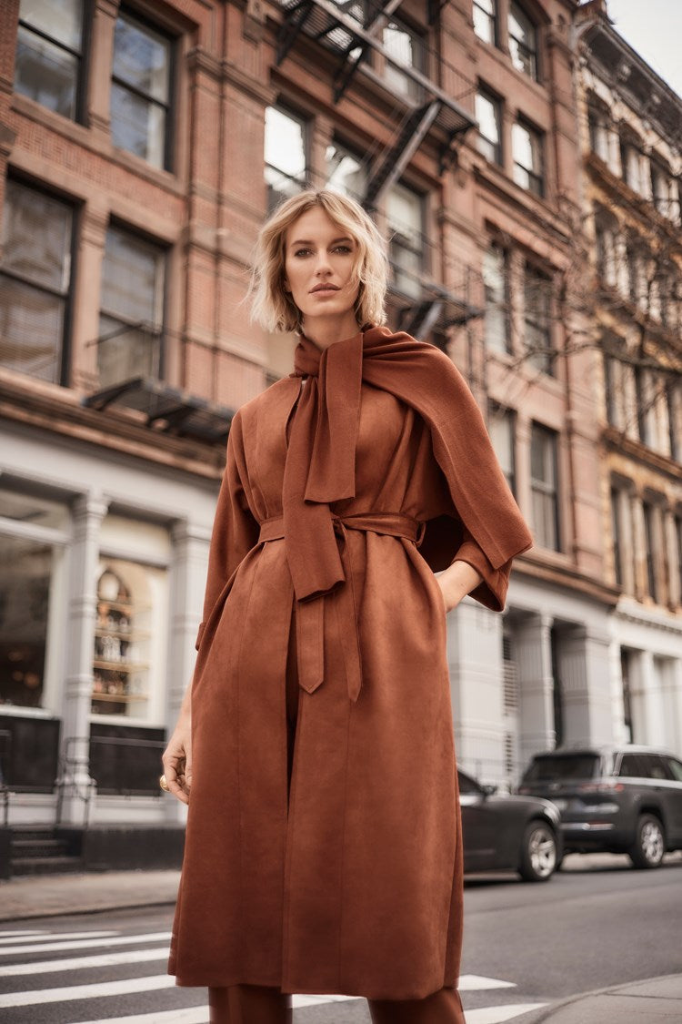 A woman stands confidently on a city street, wearing the Scuba Suede Straight Fit Coat 243037 by Joseph Ribkoff, featuring stylish three-quarter dolman sleeves and a scarf-like accessory. The urban backdrop includes brick buildings and parked cars. Her shoulder-length blonde hair is styled loosely.