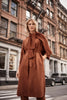 A woman stands confidently on a city street, wearing the Scuba Suede Straight Fit Coat 243037 by Joseph Ribkoff, featuring stylish three-quarter dolman sleeves and a scarf-like accessory. The urban backdrop includes brick buildings and parked cars. Her shoulder-length blonde hair is styled loosely.