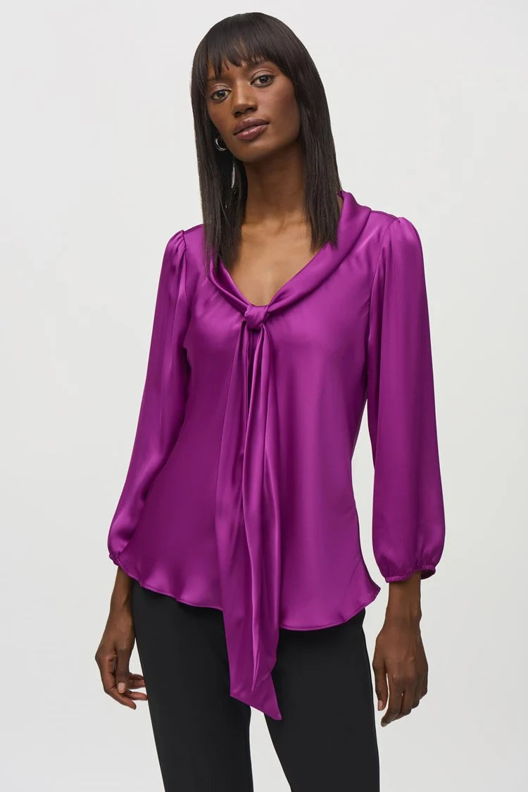 A person wearing the Joseph Ribkoff Satin Top with Bow Tie Neckline 244077 in a vibrant purple hue, featuring three-quarter sleeves and a V-shaped neckline. The blouse is paired with black pants. The person has long, straight hair and is standing against a plain, light background.