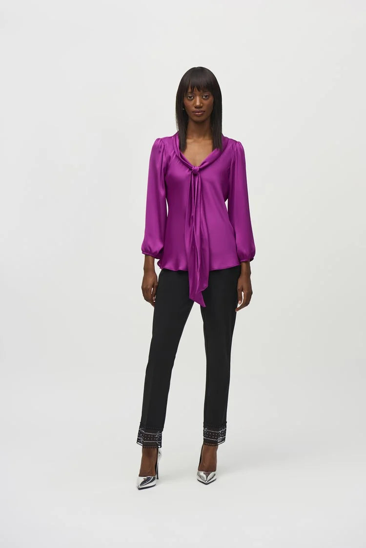 A person wearing the Joseph Ribkoff Satin Top with Bow Tie Neckline 244077 in a vibrant purple hue, featuring three-quarter sleeves and a V-shaped neckline. The blouse is paired with black pants. The person has long, straight hair and is standing against a plain, light background.