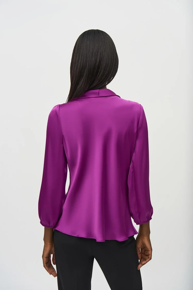 A person wearing the Joseph Ribkoff Satin Top with Bow Tie Neckline 244077 in a vibrant purple hue, featuring three-quarter sleeves and a V-shaped neckline. The blouse is paired with black pants. The person has long, straight hair and is standing against a plain, light background.