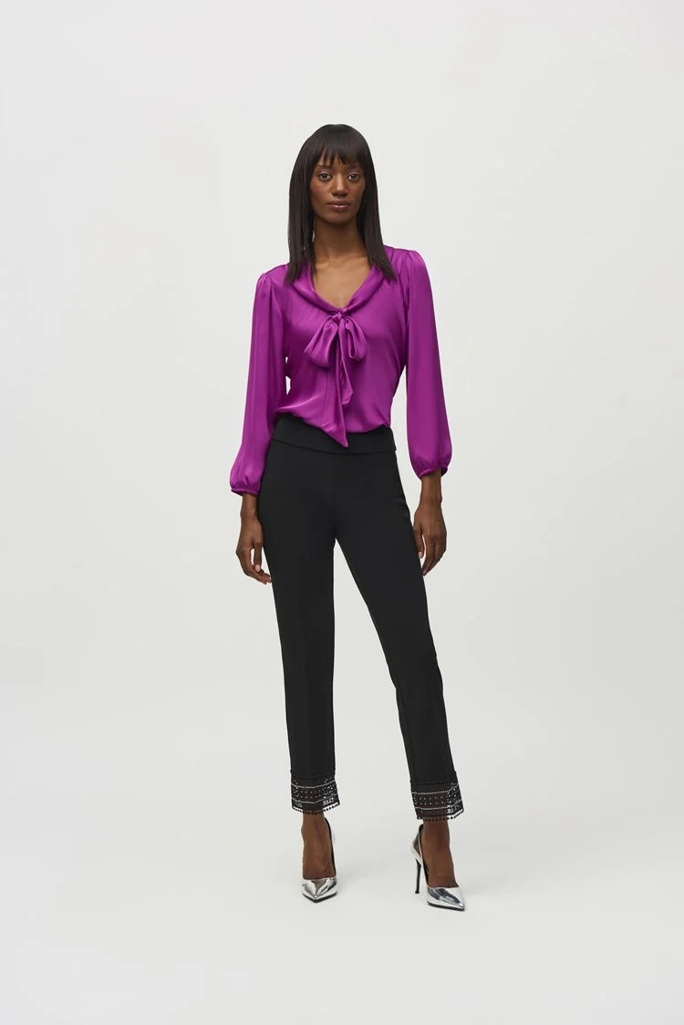 A person wearing the Joseph Ribkoff Satin Top with Bow Tie Neckline 244077 in a vibrant purple hue, featuring three-quarter sleeves and a V-shaped neckline. The blouse is paired with black pants. The person has long, straight hair and is standing against a plain, light background.