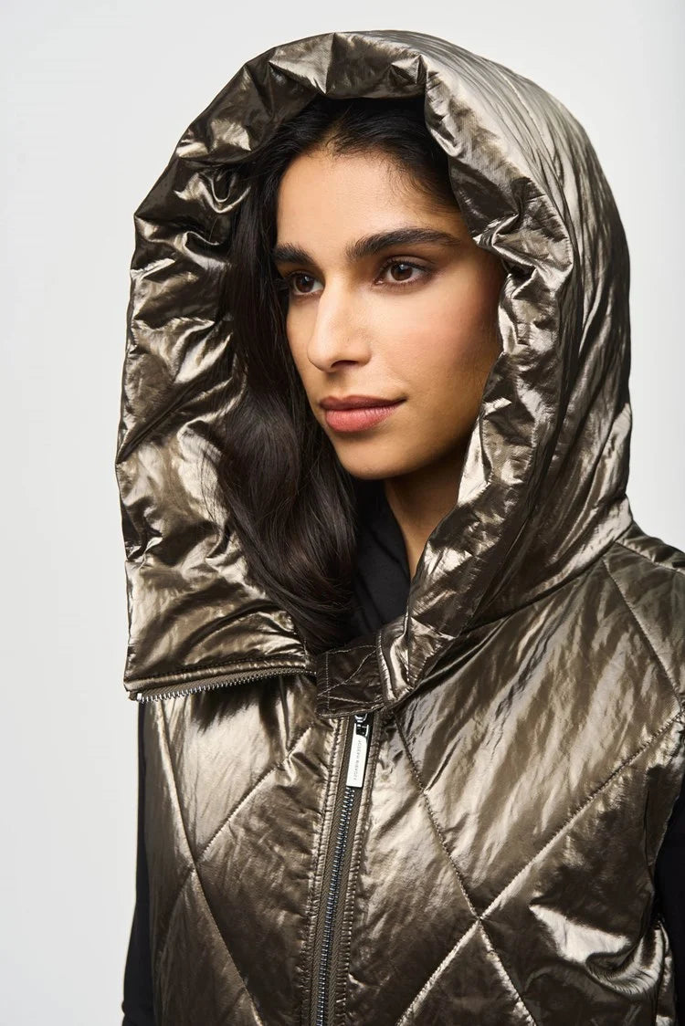 A person with long dark hair stands against a concrete wall, wearing the Joseph Ribkoff Quilted Hooded Puffer Vest over a black top and taupe pants, paired with black ankle boots. With their hands in the vest pockets and looking directly at the camera, this outfit is perfect for the cooler seasons.