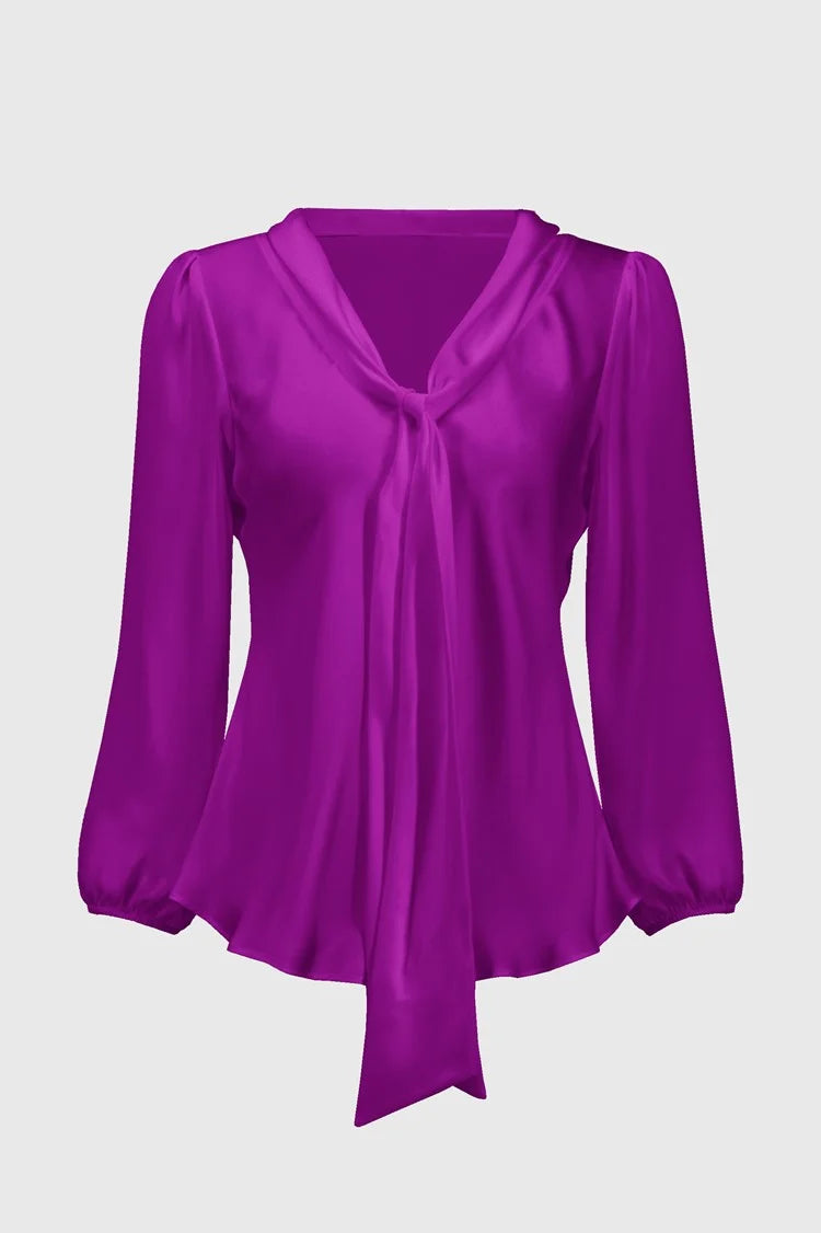 A person wearing the Joseph Ribkoff Satin Top with Bow Tie Neckline 244077 in a vibrant purple hue, featuring three-quarter sleeves and a V-shaped neckline. The blouse is paired with black pants. The person has long, straight hair and is standing against a plain, light background.