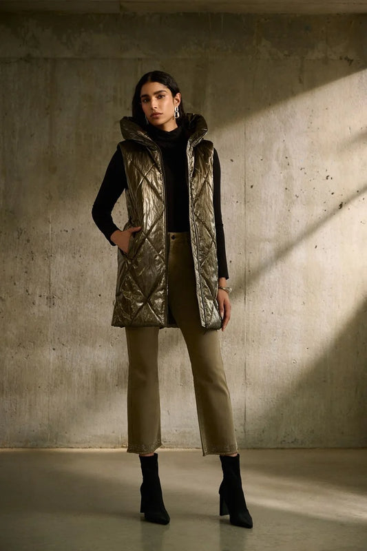 A person with long dark hair stands against a concrete wall, wearing the Joseph Ribkoff Quilted Hooded Puffer Vest over a black top and taupe pants, paired with black ankle boots. With their hands in the vest pockets and looking directly at the camera, this outfit is perfect for the cooler seasons.