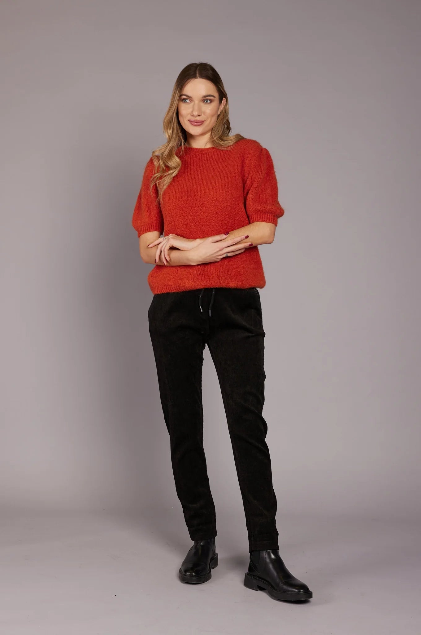 A woman stands against a plain gray background. She is wearing a red short-sleeved sweater, ELISSIA NF21600 Pants, and black boots. She has long, wavy blonde hair and is smiling slightly while looking to the side. Her hands are loosely clasped in front of her.