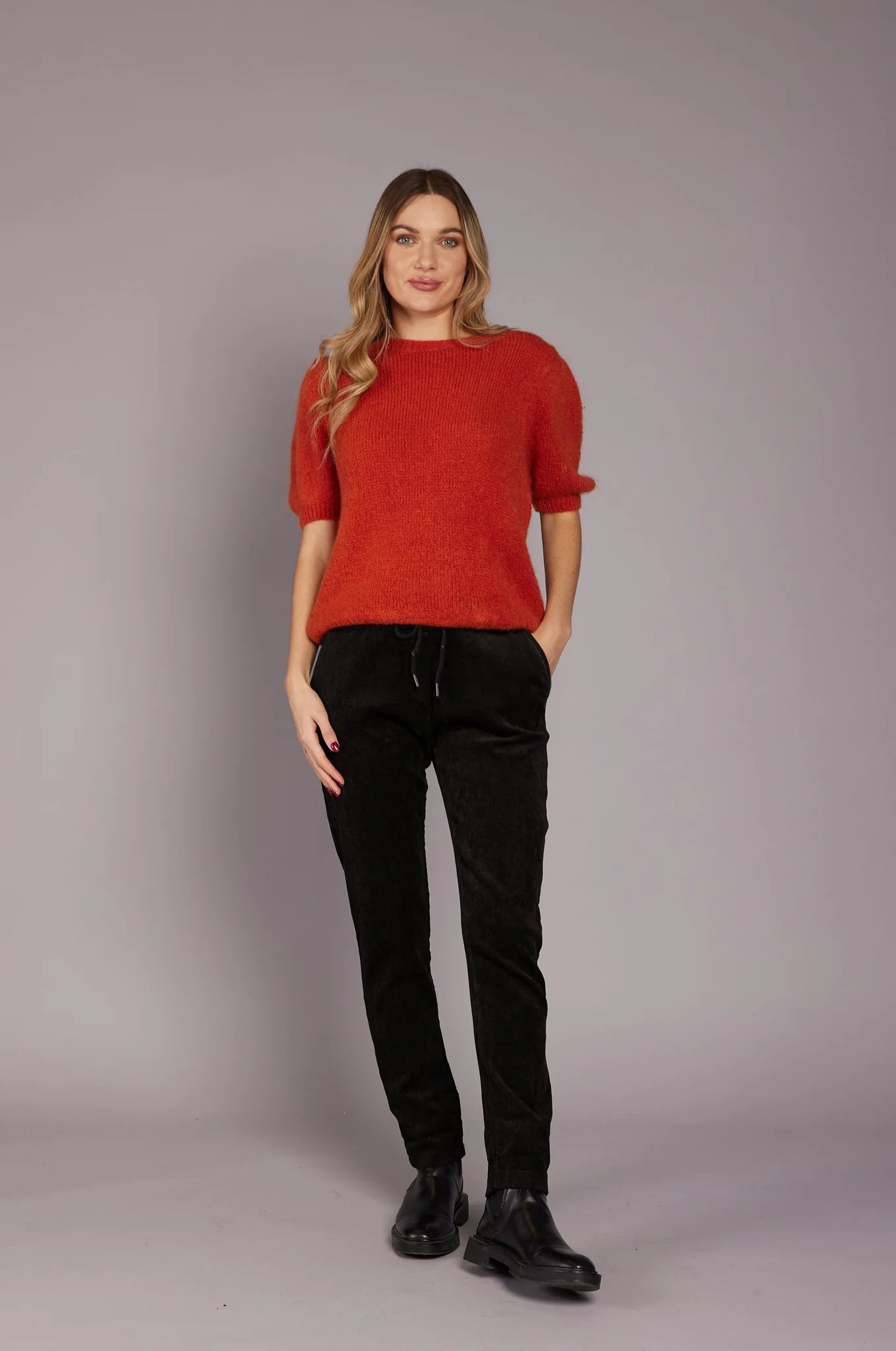 A woman stands against a plain gray background. She is wearing a red short-sleeved sweater, ELISSIA NF21600 Pants, and black boots. She has long, wavy blonde hair and is smiling slightly while looking to the side. Her hands are loosely clasped in front of her.