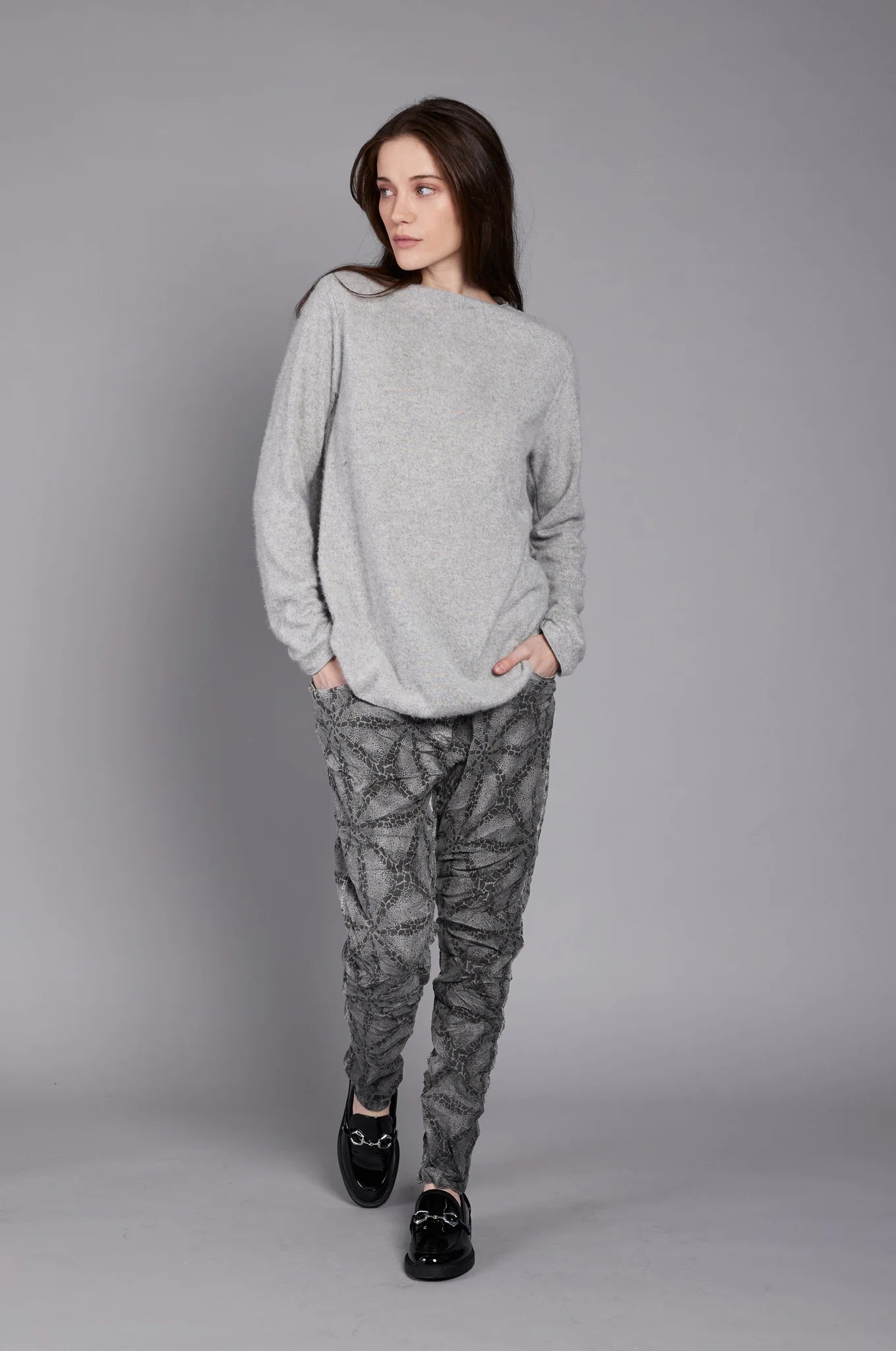 A person with long hair is standing against a plain gray background. They are wearing the JY4138 TOP by ELISSIA, made of soft breathable fabric, and patterned gray pants, with their left hand resting in their pants pocket and their right arm hanging by their side.