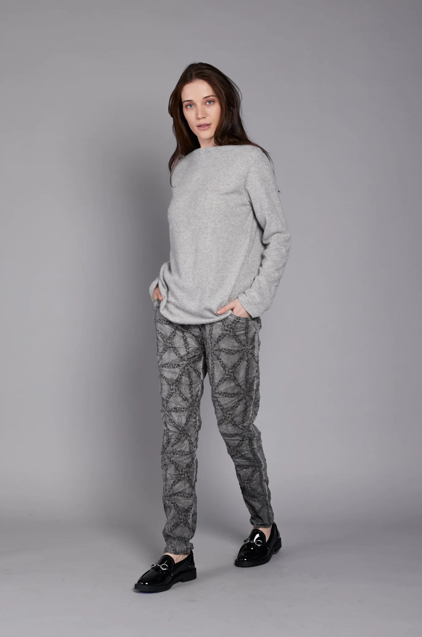 A person with long hair is standing against a plain gray background. They are wearing the JY4138 TOP by ELISSIA, made of soft breathable fabric, and patterned gray pants, with their left hand resting in their pants pocket and their right arm hanging by their side.